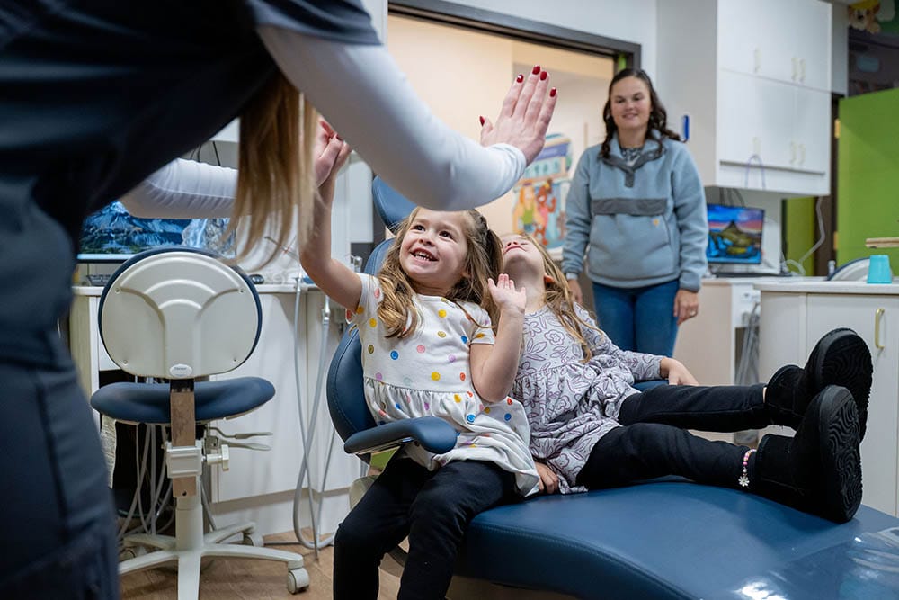 Smiling young girl in a dental chair gives a high-five to a dental professional while her parent watches in the background.