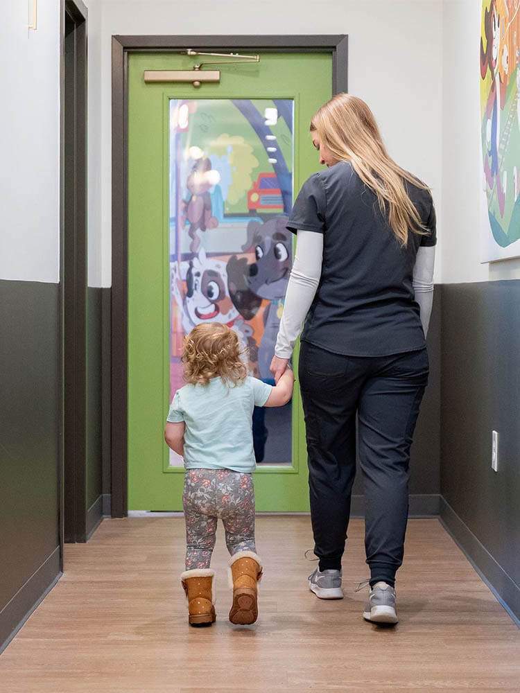 Dental staff member holds a toddler’s hand while walking down a clinic hallway toward a colorful door.