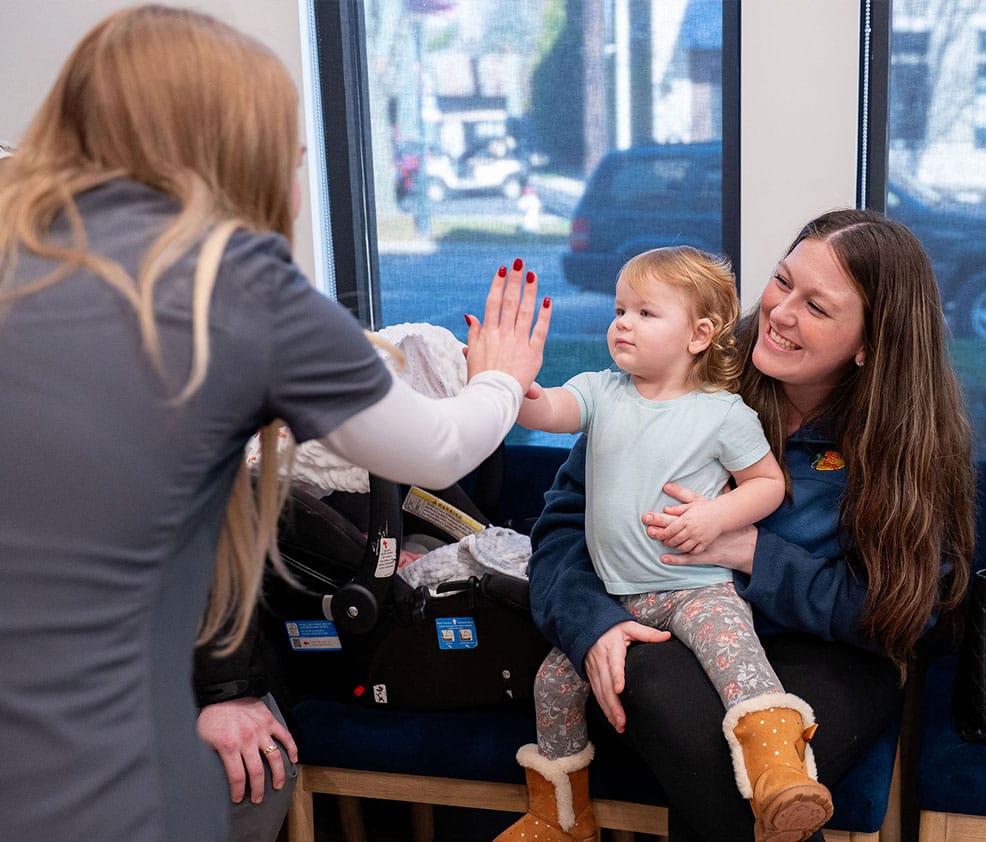 A friendly dental staff member giving a high-five to a toddler sitting on her mother's lap at Canal Kids Pediatric Dentistry in Canal Winchester.