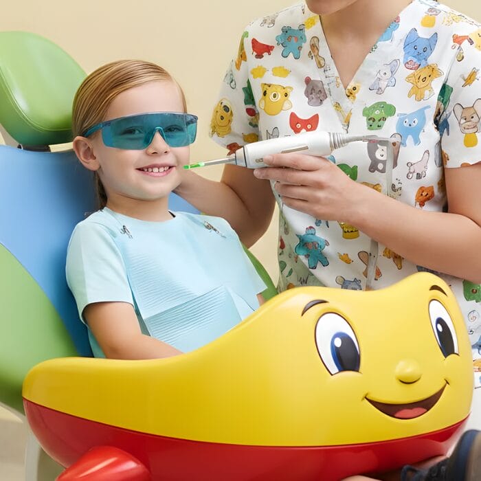 Smiling child wearing protective glasses during a pediatric laser dental treatment, seated in a colorful airplane-themed dental chair while a clinician uses a handheld laser device.